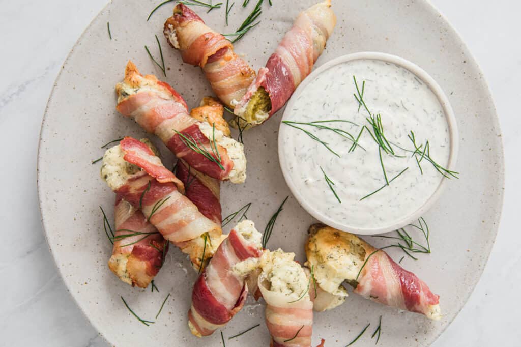 Bacon pickle boats stuffed with cream cheese on a plate next to a bowl of dip.