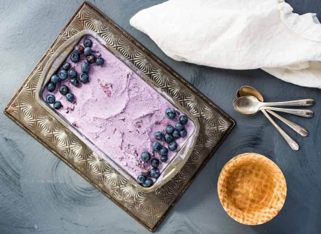 An overhead shot of homemade blueberry ice cream in a metal loaf pan next to waffle bowls, spoons, and a cream colored napkin.