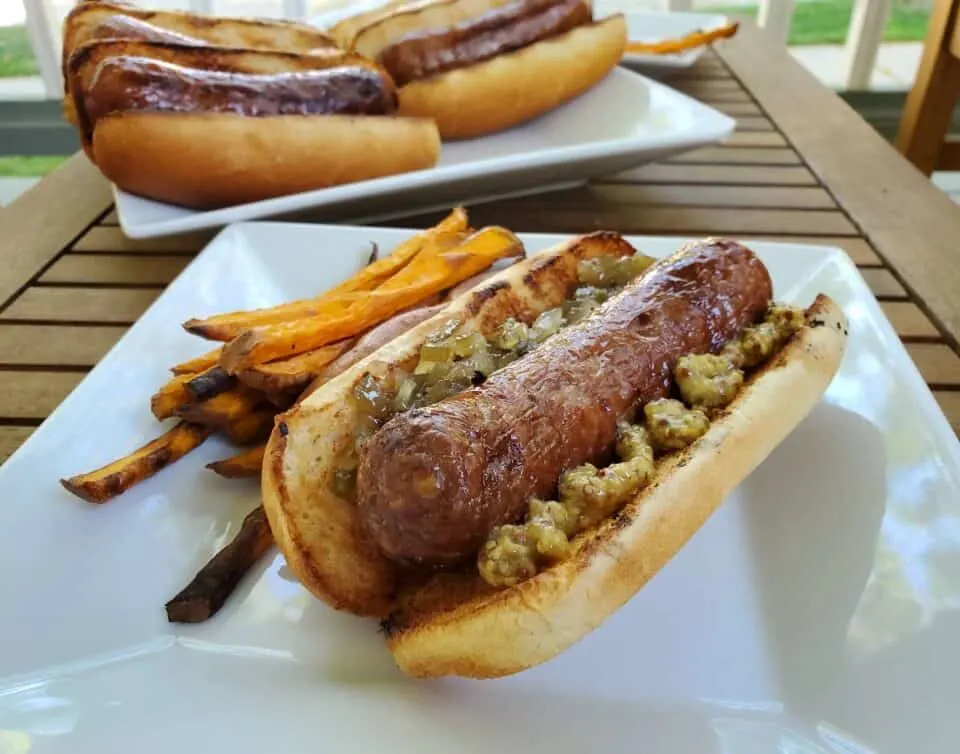 Image shows a beer boiled brat on a bun with sweet potato fries on a plate and more brats in the background.