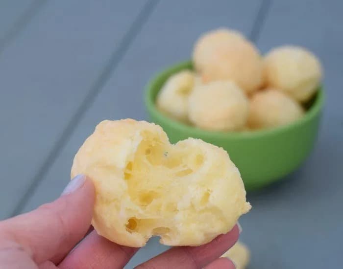 A hand holds a bite of Brazilian cheese bread broken in half with a full bowl of more pao e queijo out of focus in the background.