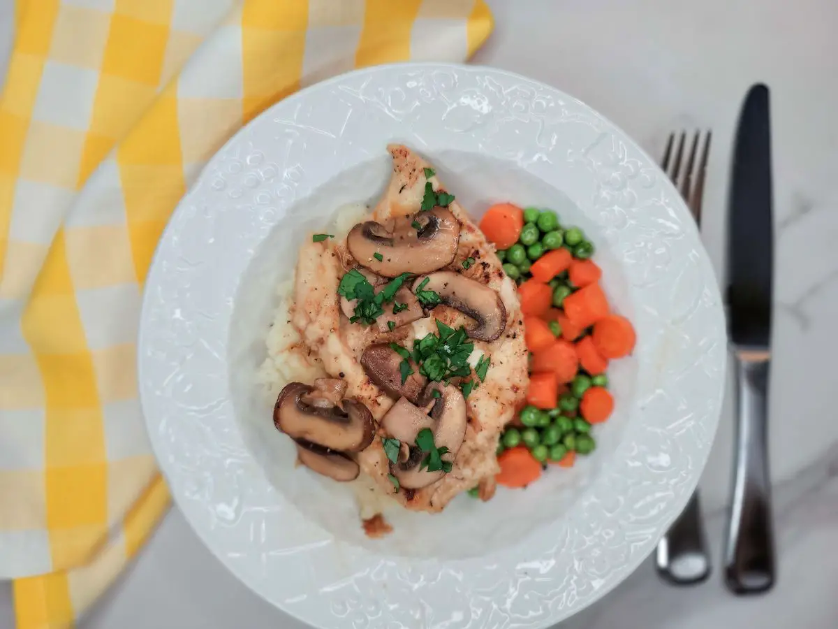 A chicken breast topped with marsala mushroom sauce atop mashed potatoes and served with buttered peas and carrots.