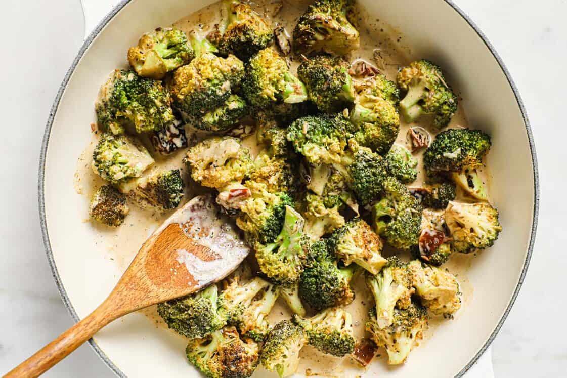 A pan of creamy broccoli with sun-dried tomatoes with a wooden spoon.