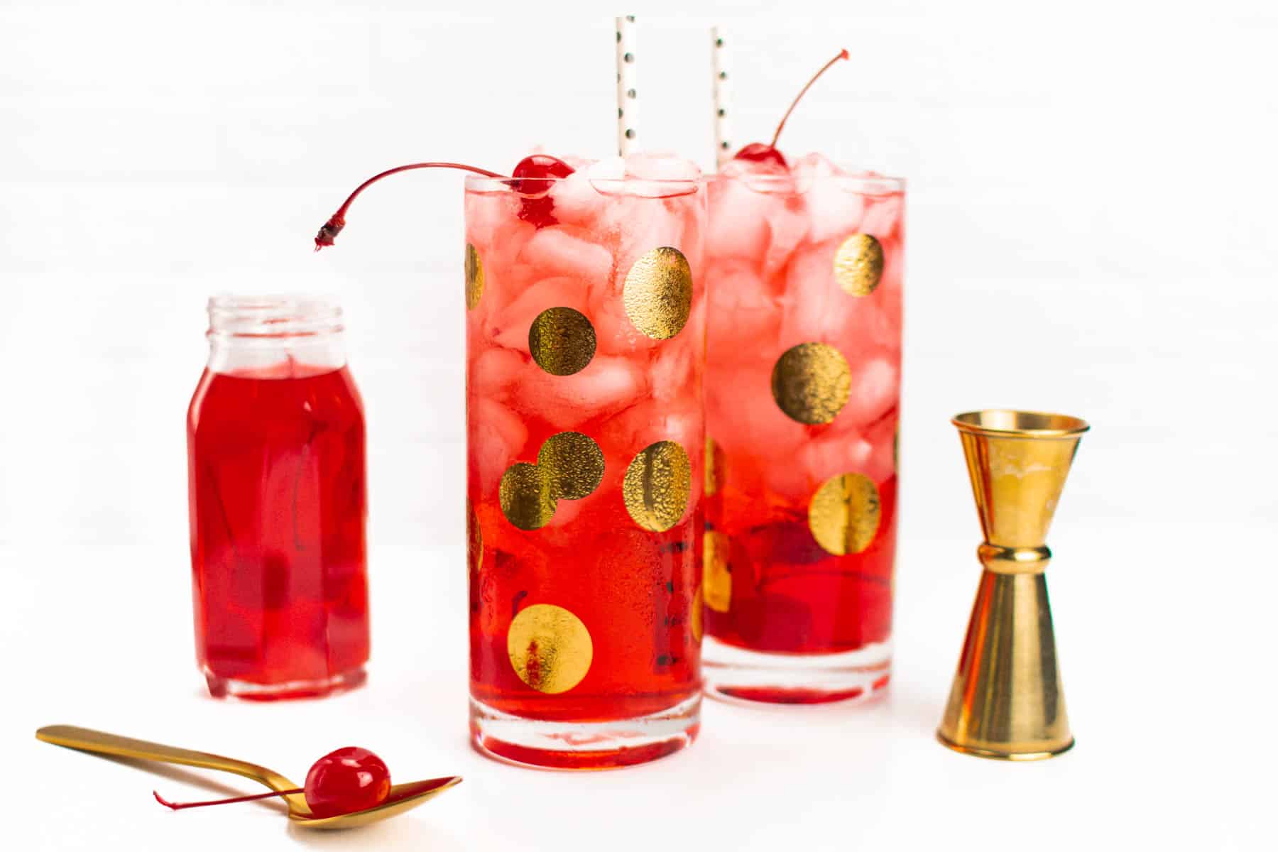Two dirty Shirley Temple cocktails with a jigger, a spoon and a jar of maraschino cherries.
