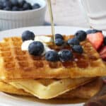 A stack of waffles with fruit and pouring syrup.