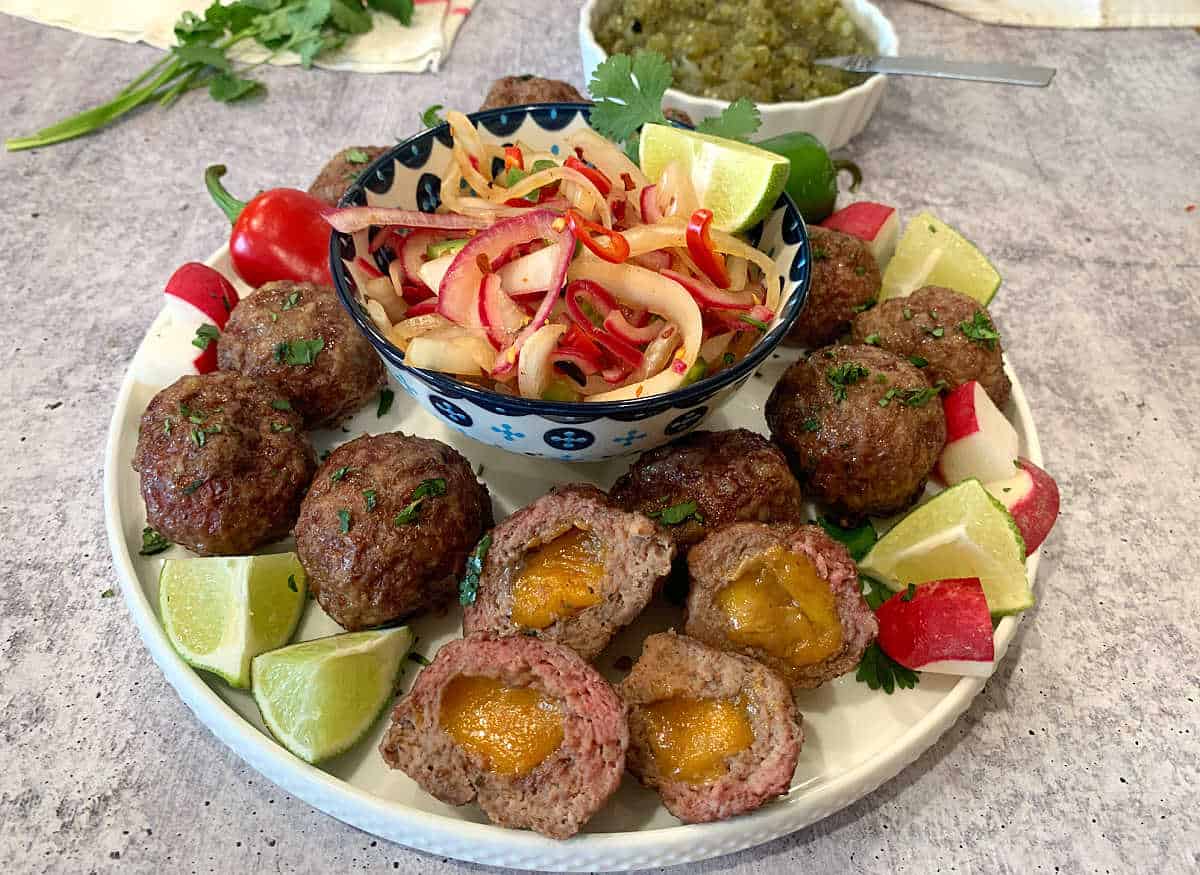 A white platter with cheese stuffed meatballs, limes, radishes and onions.