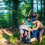 A group of five people, including two children, look at a map together while standing in a sunlit forest, planning their next adventure and discussing fun camping games.