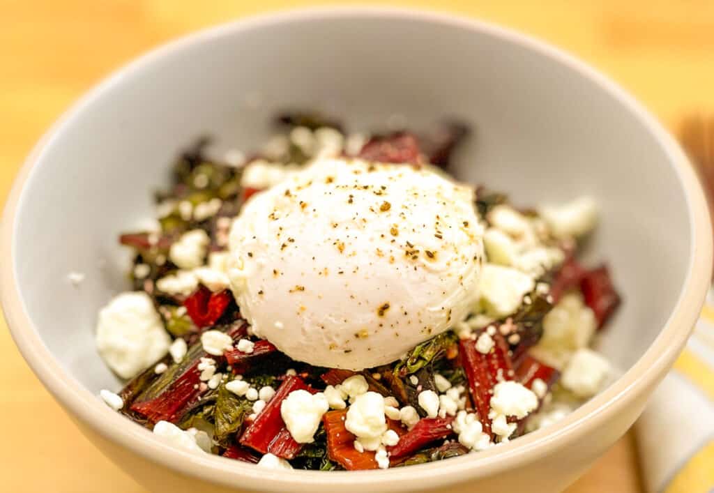 A grain bowl topped with swiss chard, goat cheese, and a poached egg in a white bowl.