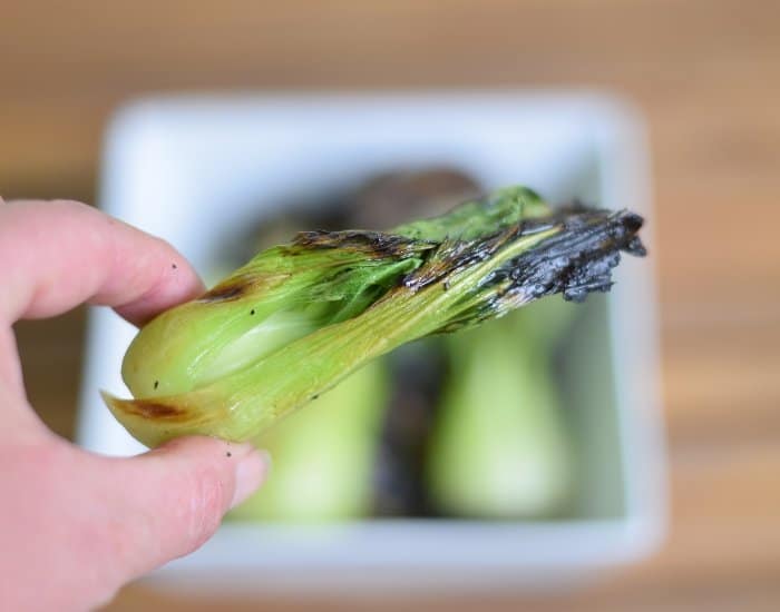 Image shows a hand holding a piece of grilled baby bok choy over a bowl containing more bok choy.
