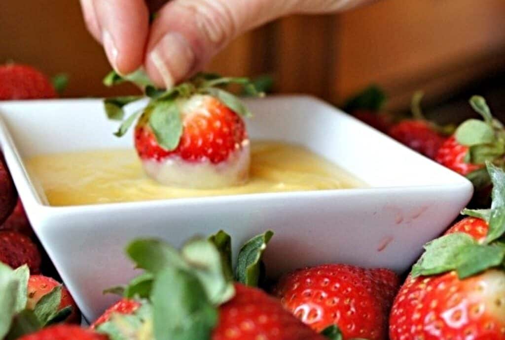 Hand dipping a strawberry into pineapple curd in a white bowl.