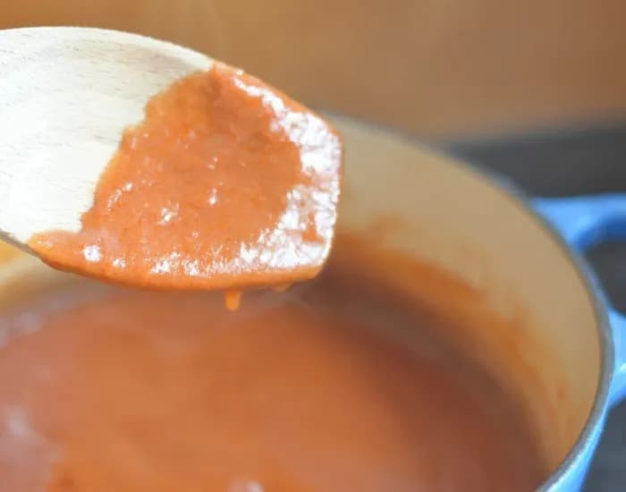 A wooden spoon has red enchilada sauce dripping off it into a blue pot filled with the remainder of the homemade enchilada sauce.