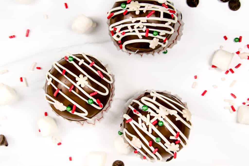 Three hot chocolate bombs, sprinkles and marshmallows on a marble background.