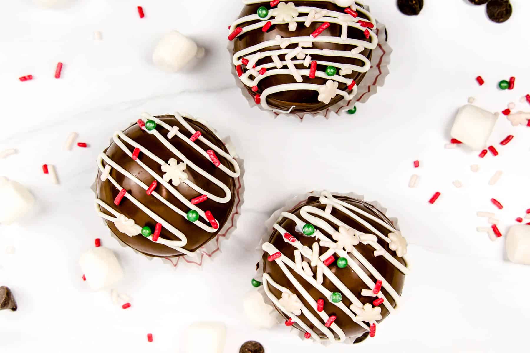 Three hot chocolate bombs, sprinkles and marshmallows on a marble background.