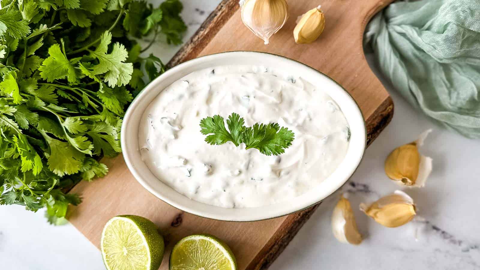 Cilantro lime crema in a white and green dish surrounded by lime halves, fresh cilantro, and garlic cloves.