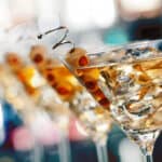 Several martini glasses filled with a brownish martini cocktail, each garnished with green olives on skewers and ice cubes, are arranged in a row with a blurred background.