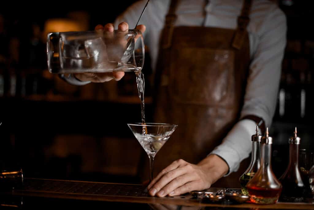 Professional bartender pouring a transparent alcoholic drink from the measuring cup to the martini glass on the bar counter in the dark blurred background