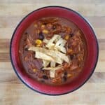 Overhead shot of chicken tortilla soup in a red bowl on wooden table with tortilla strips on the soup.