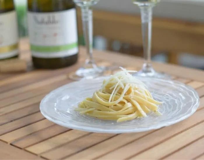 A round plate holds a serving of cacio e pepe topped with more paremsan cheese on a wooden table with wine bottles in the background.