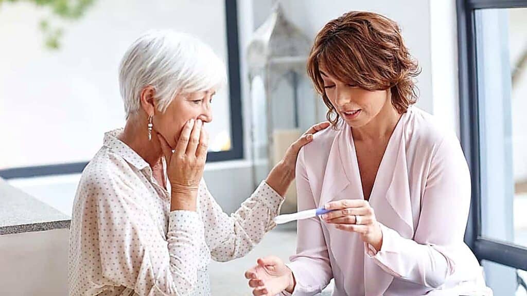Two women sit together indoors. One woman holds a pregnancy test and looks at it, while the other touches her shoulder and covers her mouth in apparent surprise, as if reacting to unexpected news—capturing a heartfelt moment that could inspire ideas on how to reveal pregnancy.