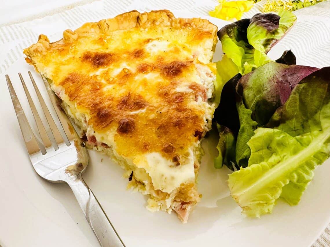 Slice of Quiche Lorraine on white plate next to fork and green salad.
