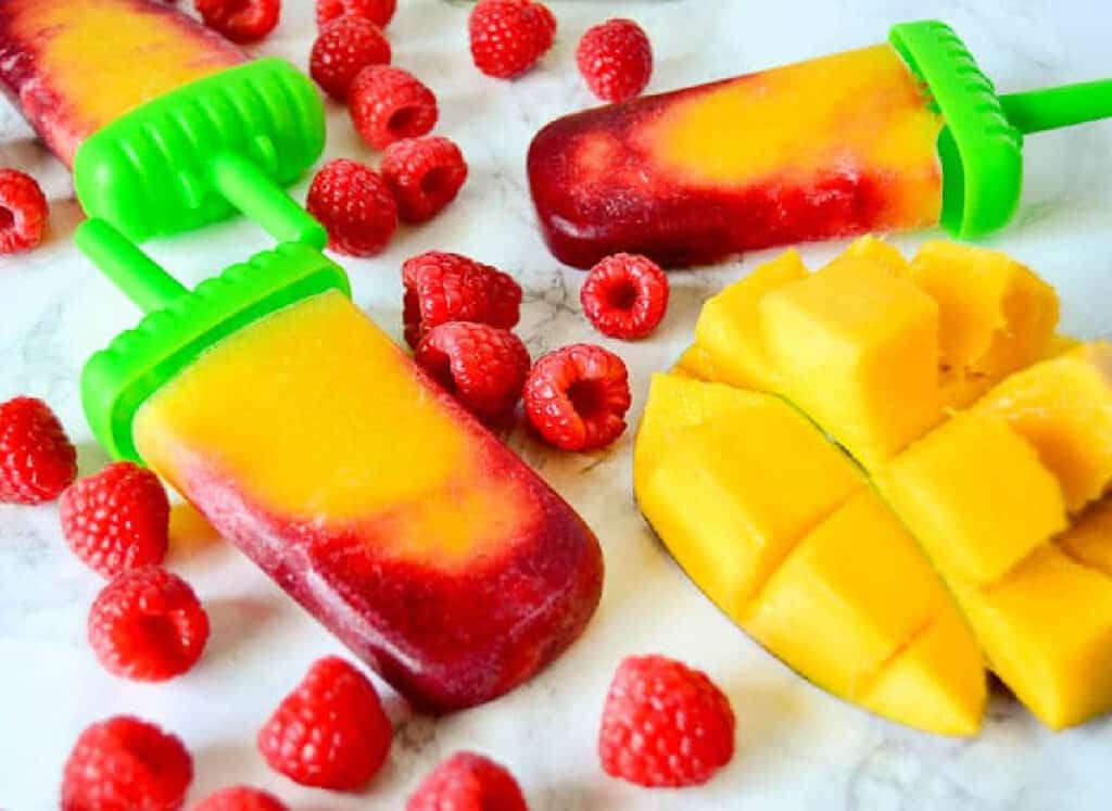 Raspberry mango popsicle on a marble background next to raspberries and mango.
