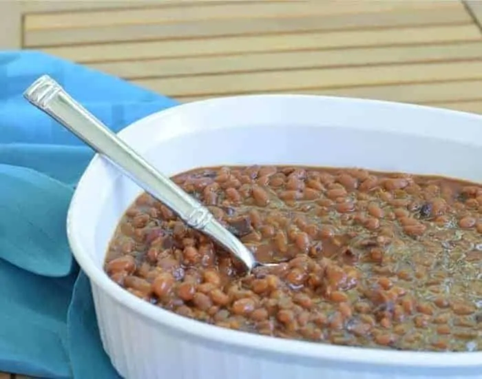 Image shows a white casserole dish filled with homemade baked beans on wooden table with a silver spoon sticking out from it.