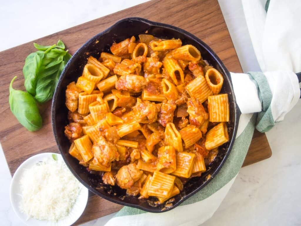 Cast iron skillet with chicken and rigatoni on a wooden cutting board next to some basil and Parmesan.