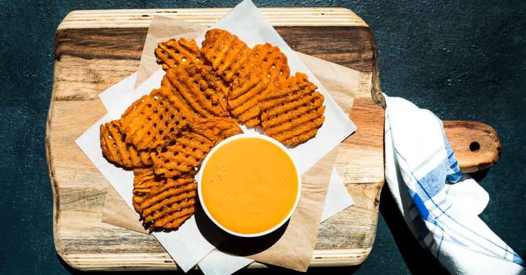 An overhead shot of spicy sweet sriracha sauce in a white ramekin next to a pile of sweet potato waffle fries on a wooden cutting board.