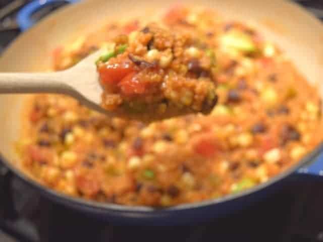 Image shows a wooden spoon holding a scoop of Mexican skillet quinoa with the full pot behind it.