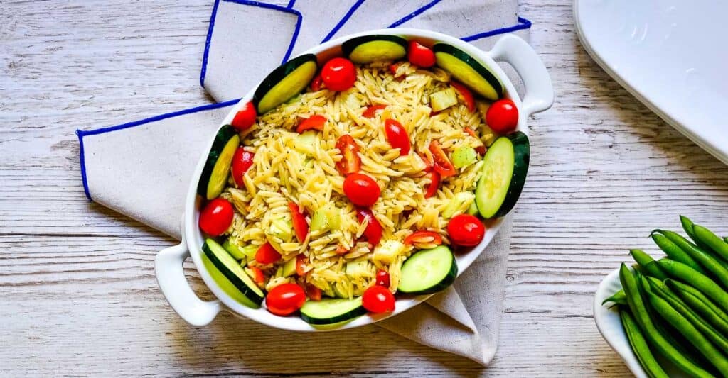 Summer orzo pasta salad in a while serving bowl on a whitewashed background next to a bowl of green beans.