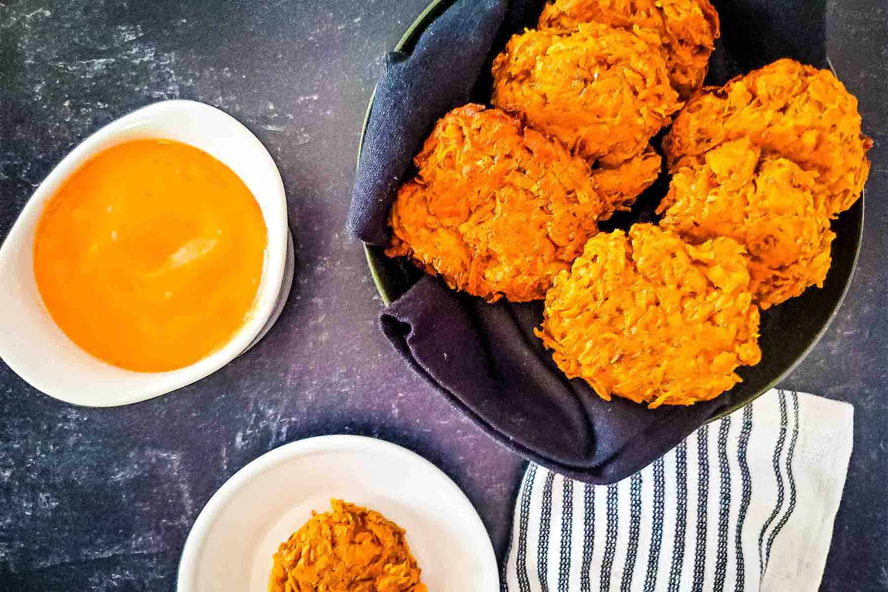 A serving basket of sweet potato fritters next to a bowl of dipping sauce and a single serving on a plate.