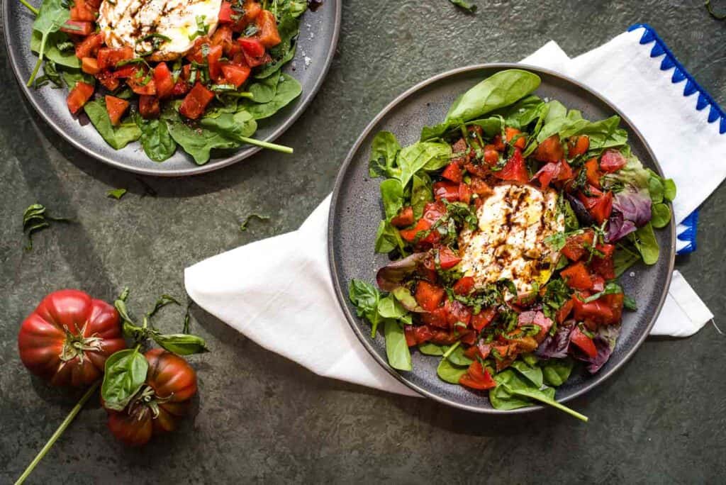 Tomato burrata salad on a grey plate next to fresh tomatoes on a grey background.