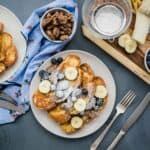 A serving of vanilla french toast next to a serving plate and bowls of candied pecans, blueberries, powdered sugar, and a banana.