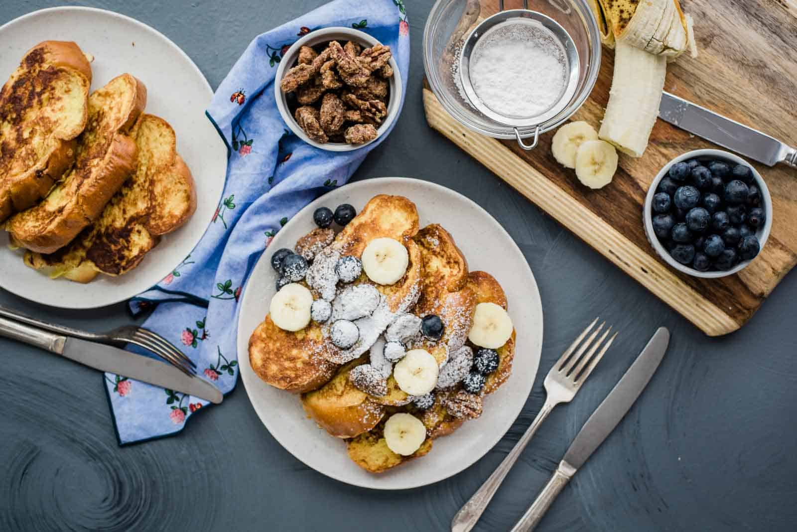 A serving of vanilla french toast next to a serving plate and bowls of candied pecans, blueberries, powdered sugar, and a banana.