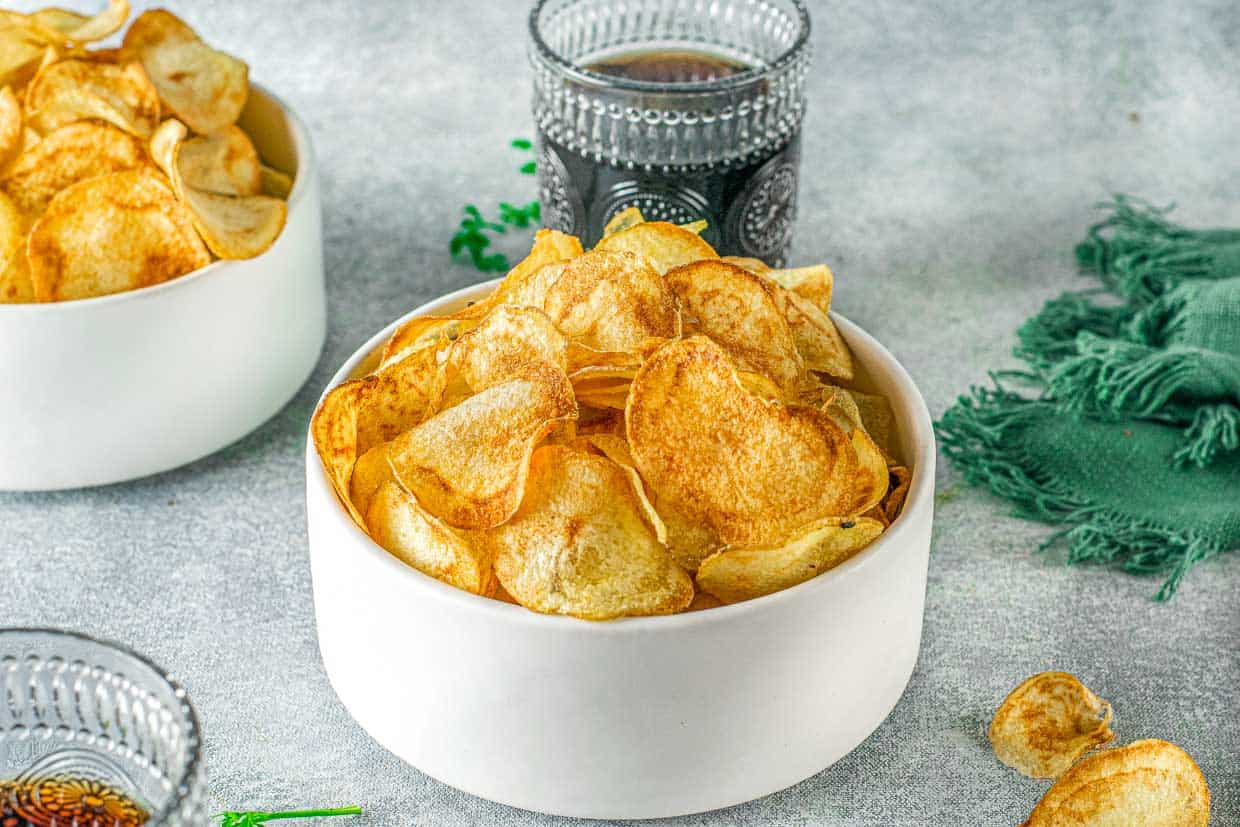 Bowl of air fryer potato chips on a white background with a soda behind it.