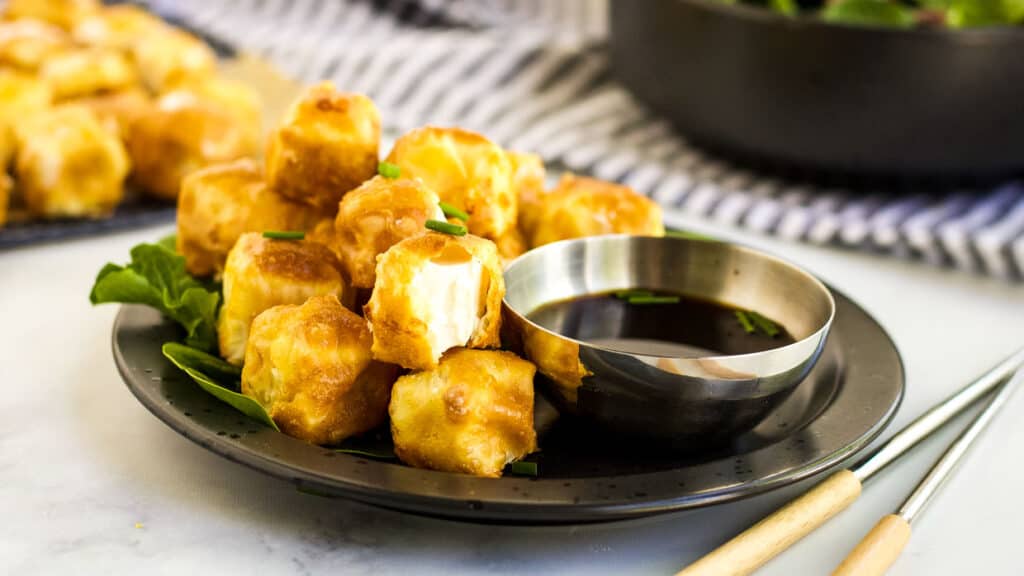 Fried tofu on a plate with one bitten into and a bowl of dipping sauce.