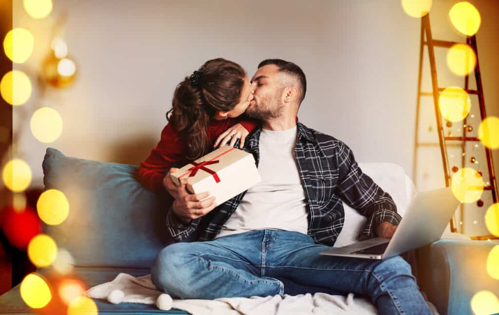 A young girl kisses a man on the cheek while giving him a wrapped present—one of the perfect anniversary gifts for him. The man sits on a sofa with his laptop, holiday lights blurred warmly in the foreground.