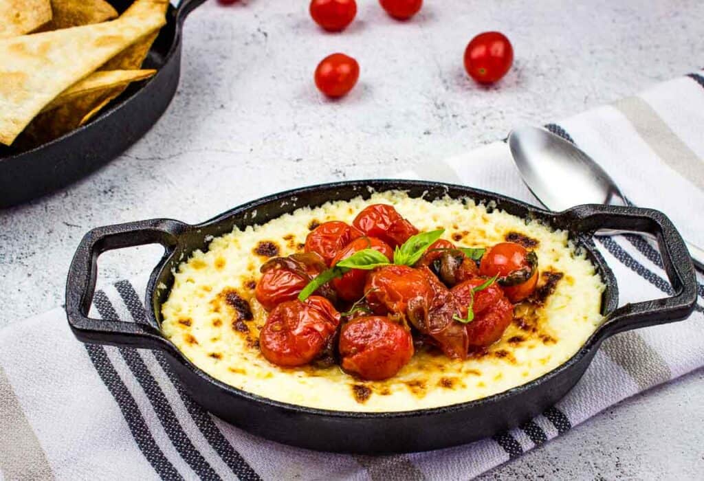 Baked Ricotta Dip with Marinated Tomatoes in a black dish with dippers in the background.