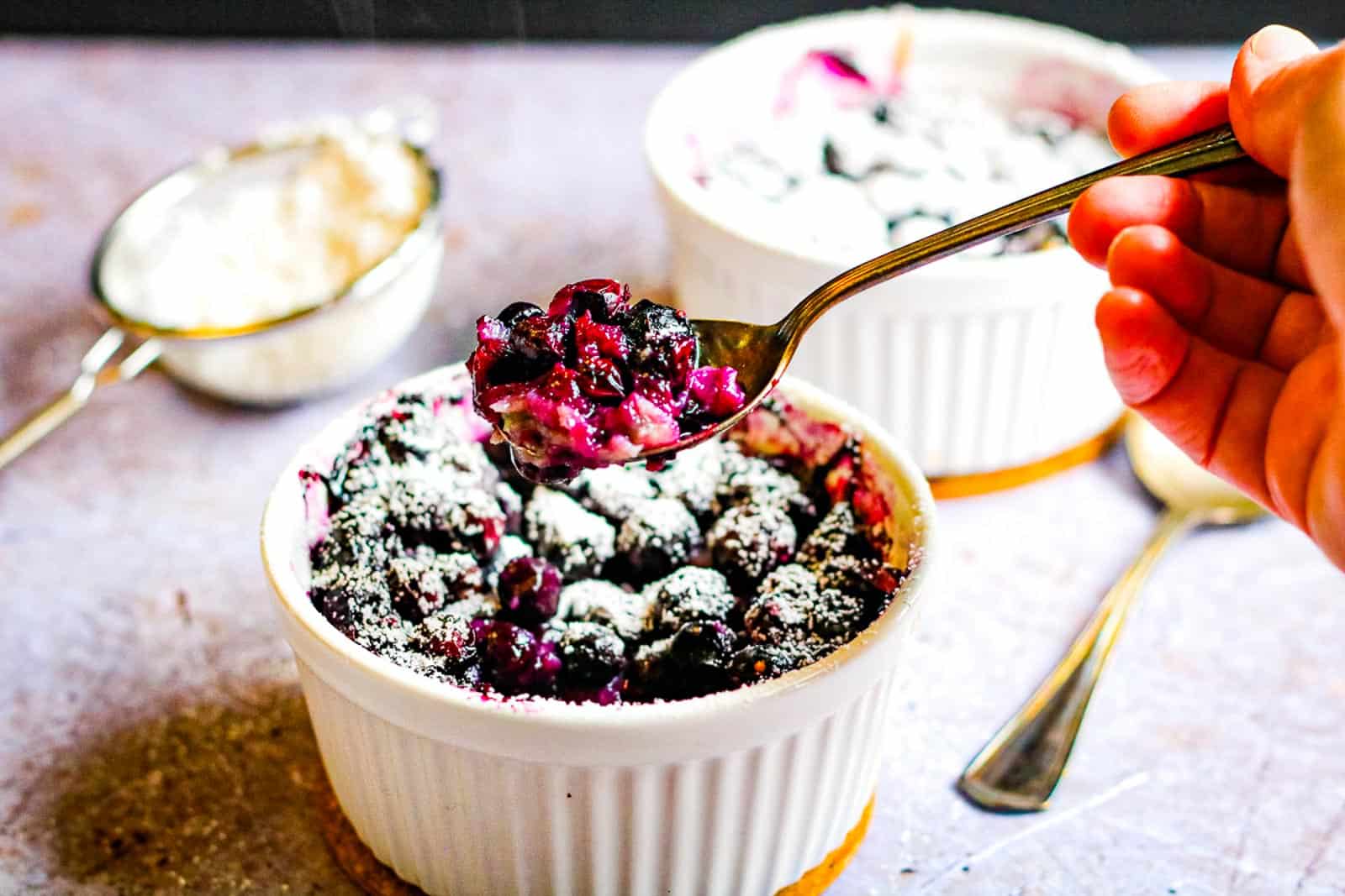 low angle shot of a white ramekin filled with blueberry clafoutis and a hand lifting a spoonful.