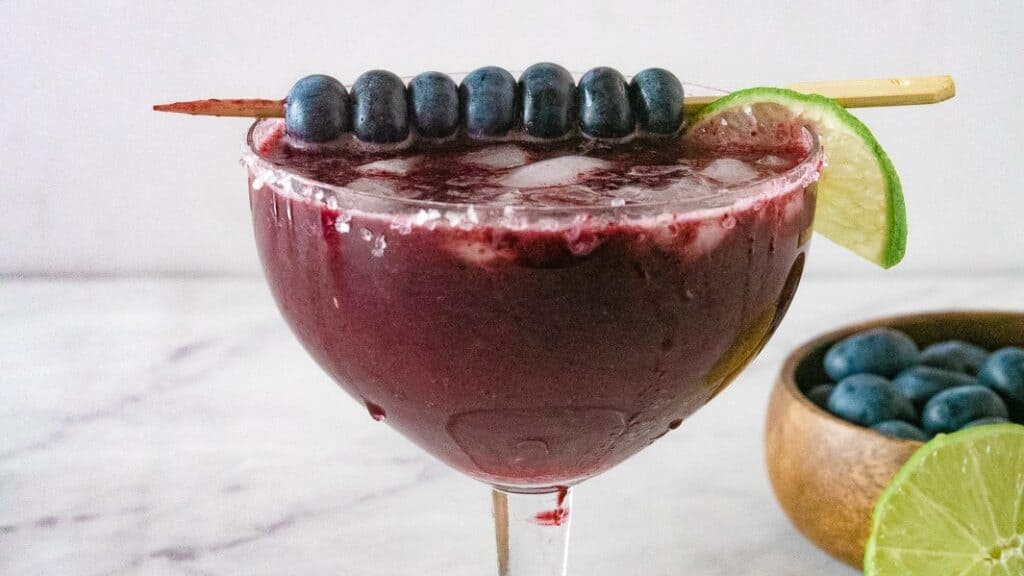Margarita glass with blueberry margarita topped with lime and a skewer of blueberries.