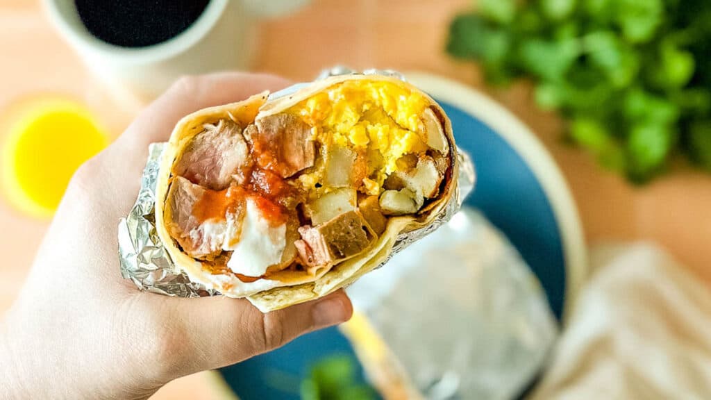 A carne asada breakfast burrito is held above a blue plate surrounded by coffee, cilantro, and orange juice.