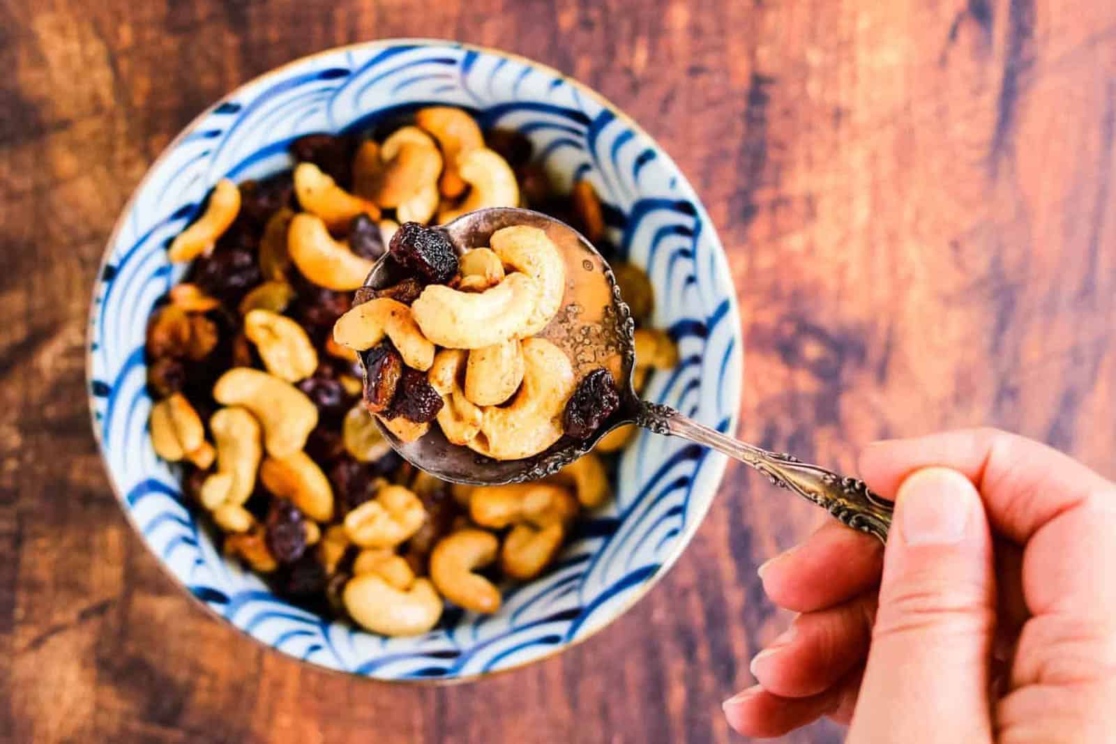 Caramelized cashews and raisins in a bowl with a silver spoon being lifted.