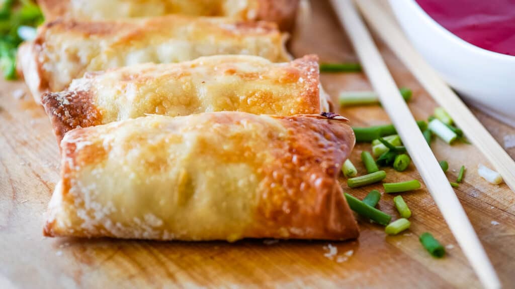 Sweet and sour chicken egg rolls on a wooden cutting board with chopsticks and green onions.