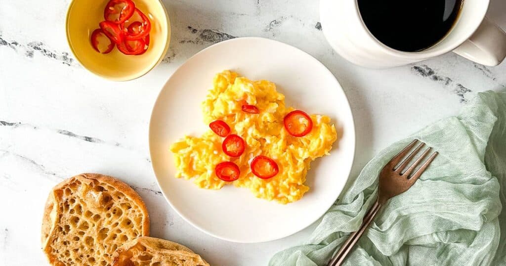 Chilli scrambled eggs on a white plate surrounded by a cup of coffee, English muffins, and a copper fork.