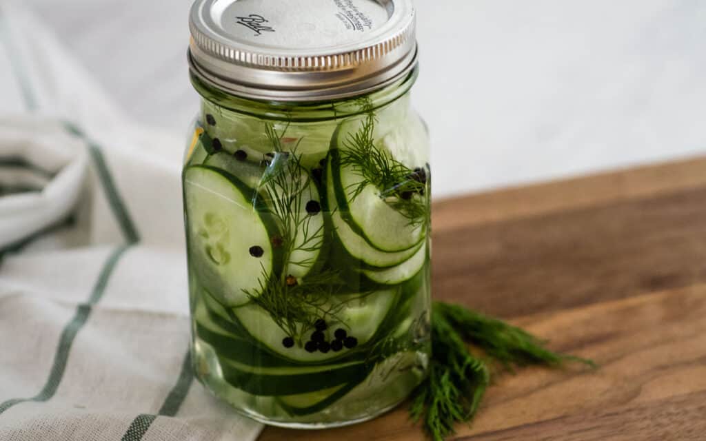 Jar of cucumber pickles with dill on a cutting board with a towel in the background.