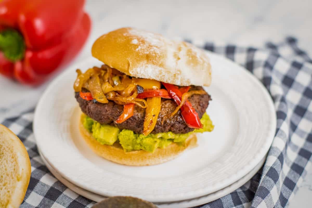 A burger topped with peppers and onions with guacamole.