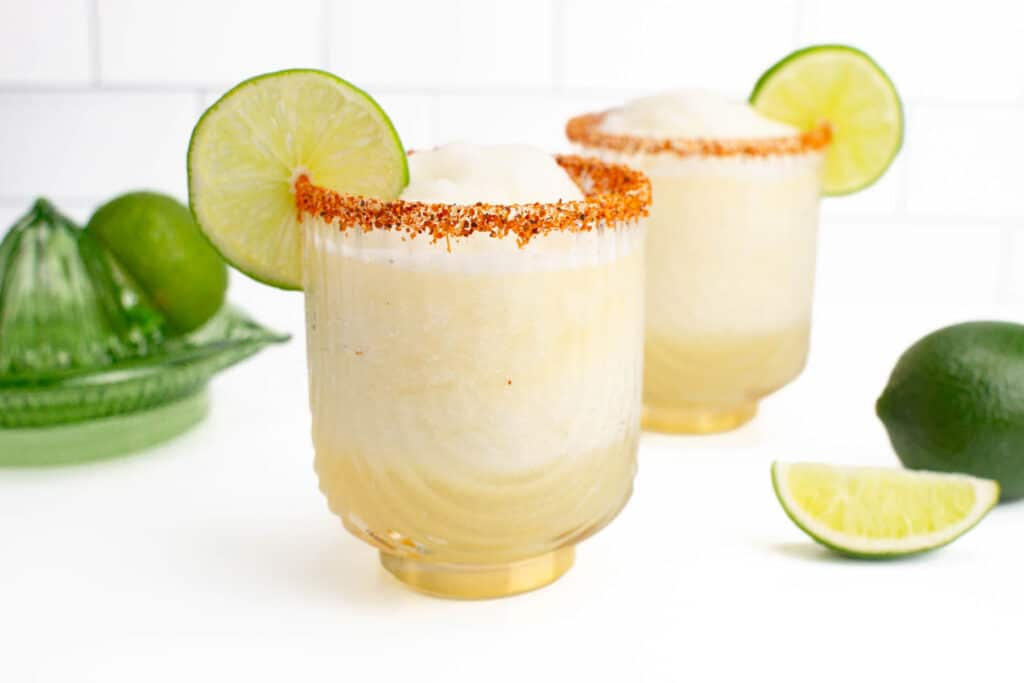 two frozen lime margaritas with chili lime salt on the rim sitting on a white countertop.