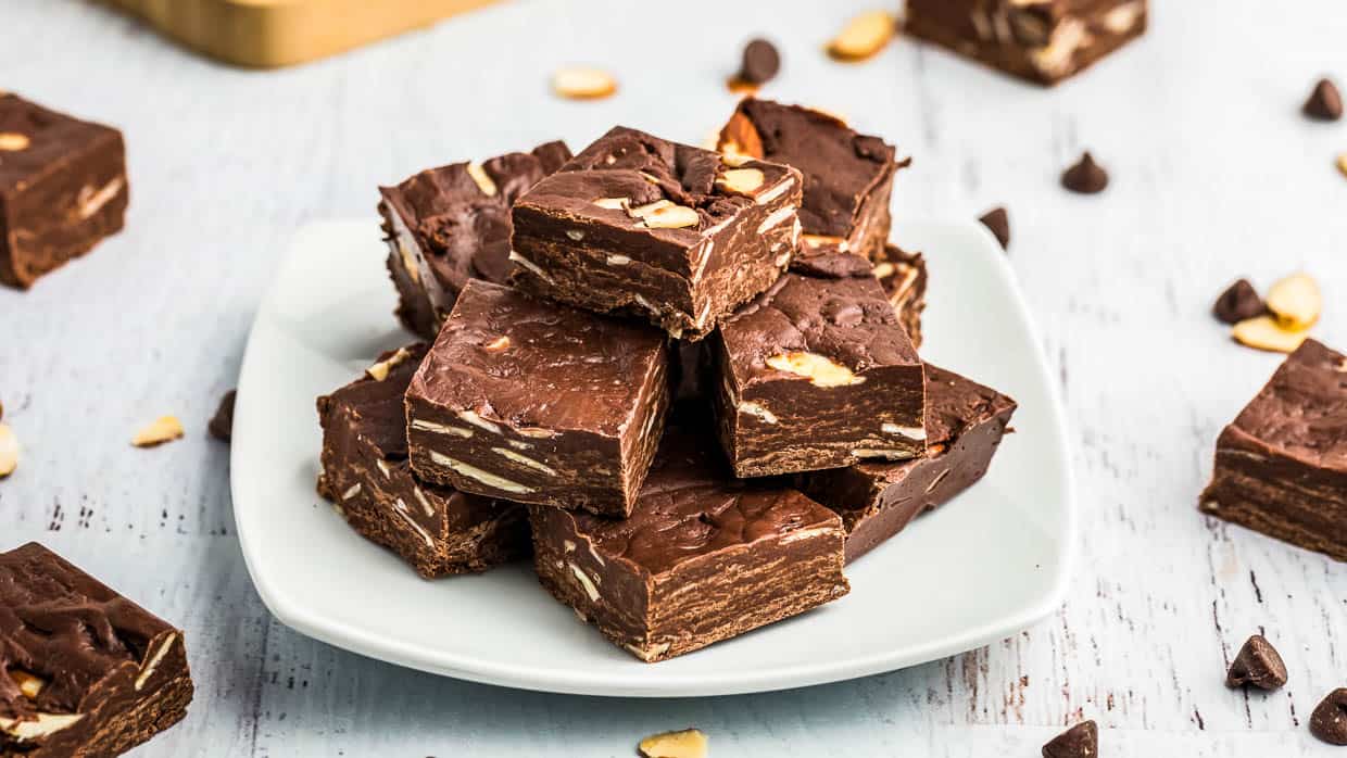 Plate of chocolate almond fudge stacked with chocolate chips and almonds scattered around.