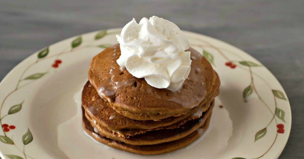 Stack of gingerbread pancakes on a plate with syrup and whipped cream.