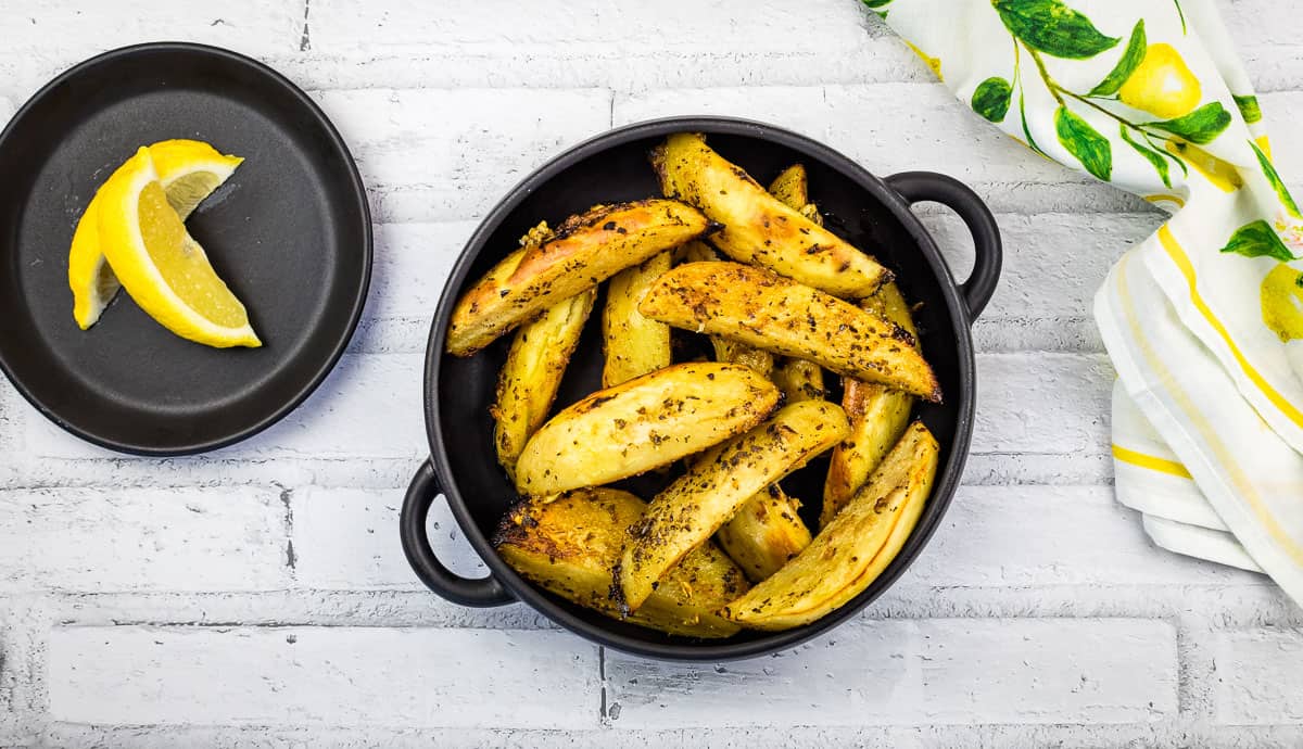 Round black dish filled with seasoned roasted potato wedges, placed on a white wooden surface alongside a white and yellow striped cloth napkin.
