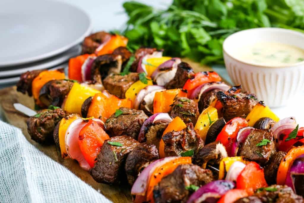 steak kabobs with peppers and onions on a cutting board with sauce and plates in the background.